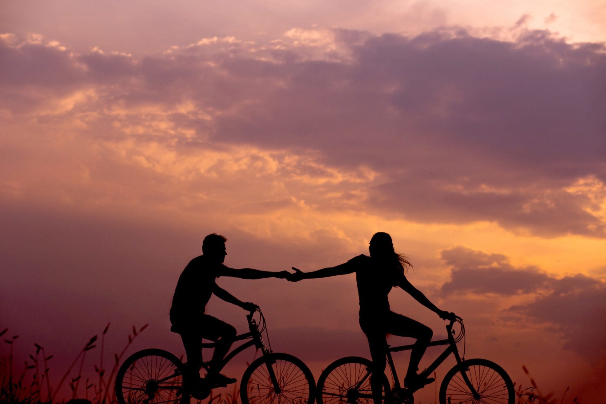young couple biking in a sunset holding hands